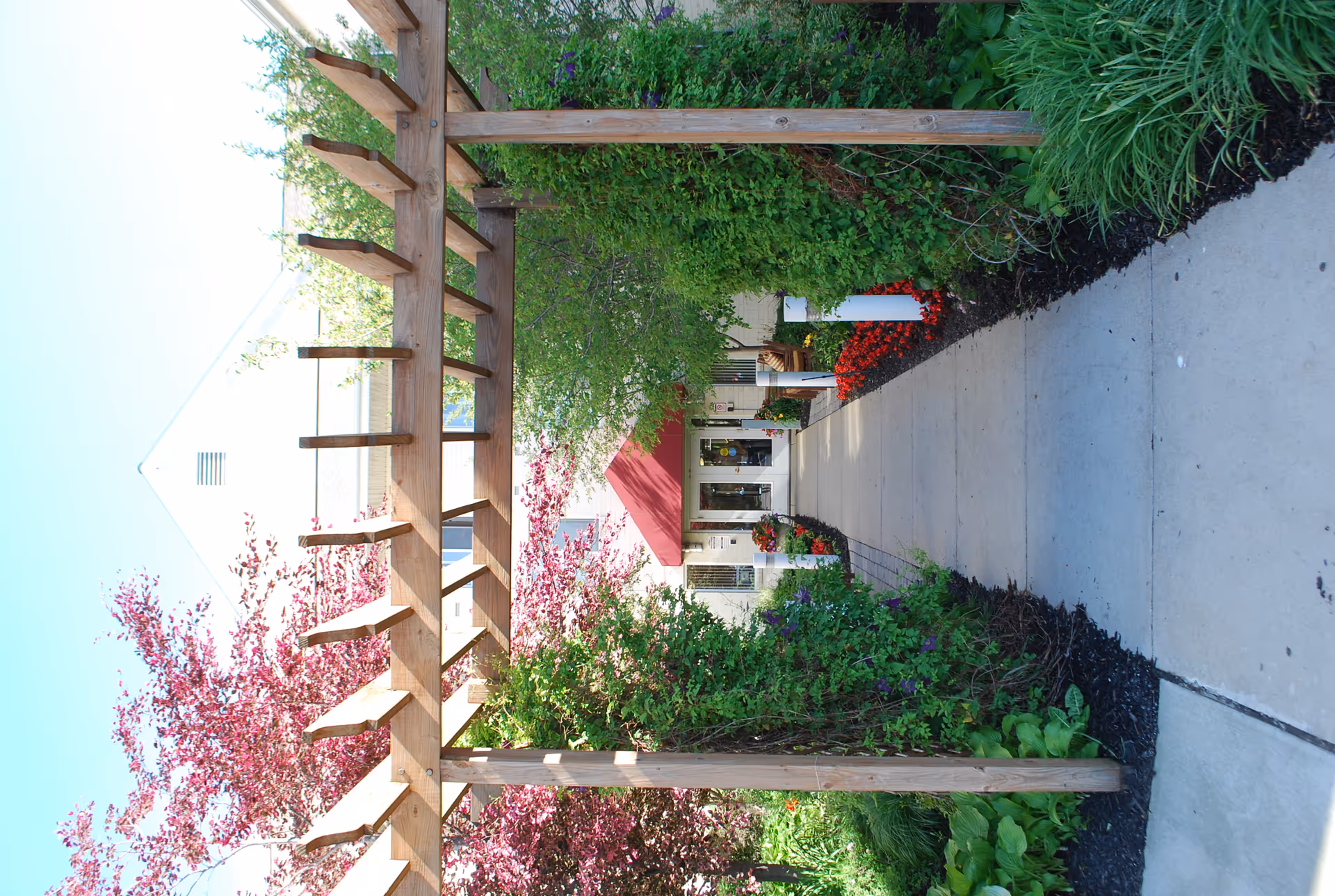 Exterior walkway with a wooden pergola guiding to the entrance, surrounded by colorful flowers.