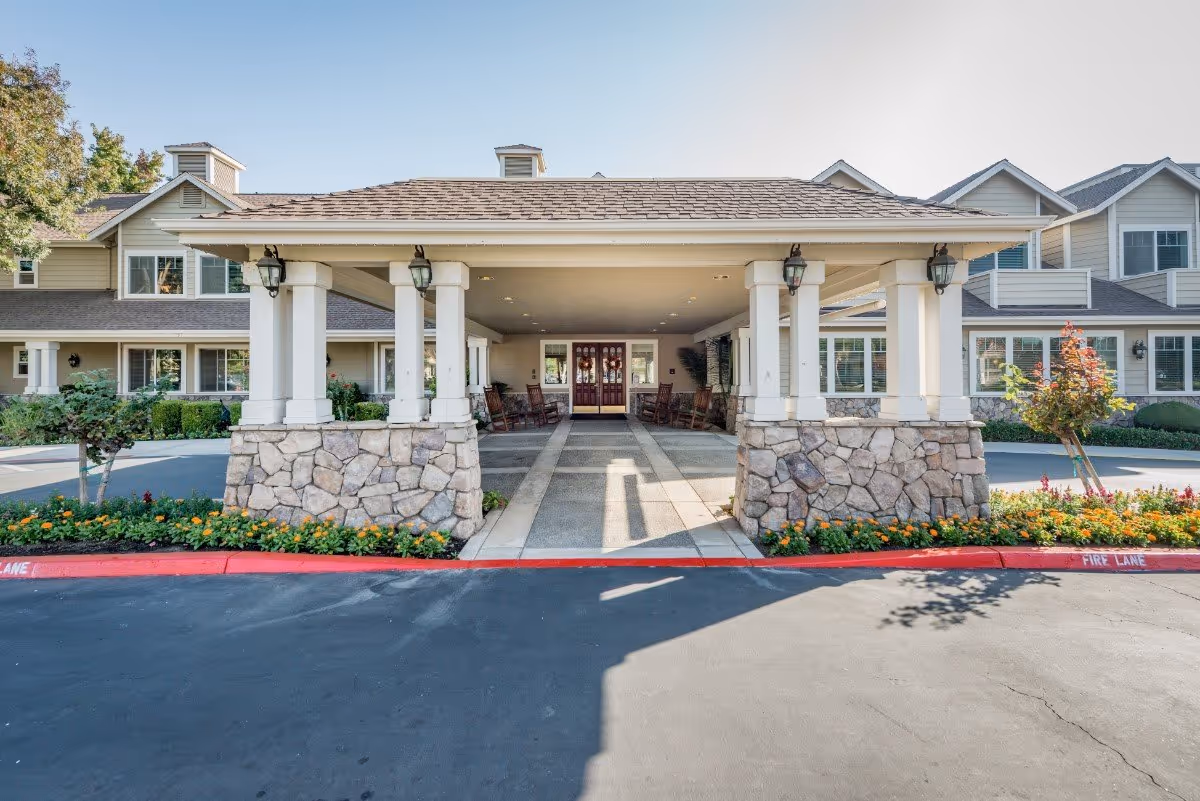 Front entrance of a senior living facility with a covered driveway supported by stone pillars and white columns. The building has beige siding, multiple windows, and a shingled roof. There are flower beds with orange and yellow flowers along the curb marked as a fire lane.