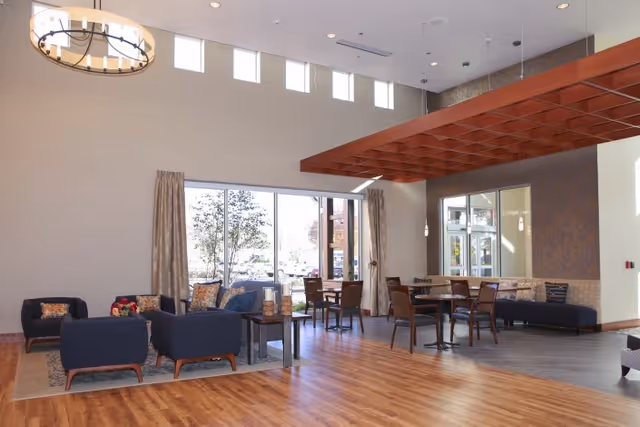 A spacious and well-lit common area with wooden flooring, featuring a seating arrangement of dark blue armchairs with colorful cushions around a small table on a rug, and several dining tables with chairs near large windows letting in natural light. The ceiling has a decorative wooden grid structure and a modern chandelier.