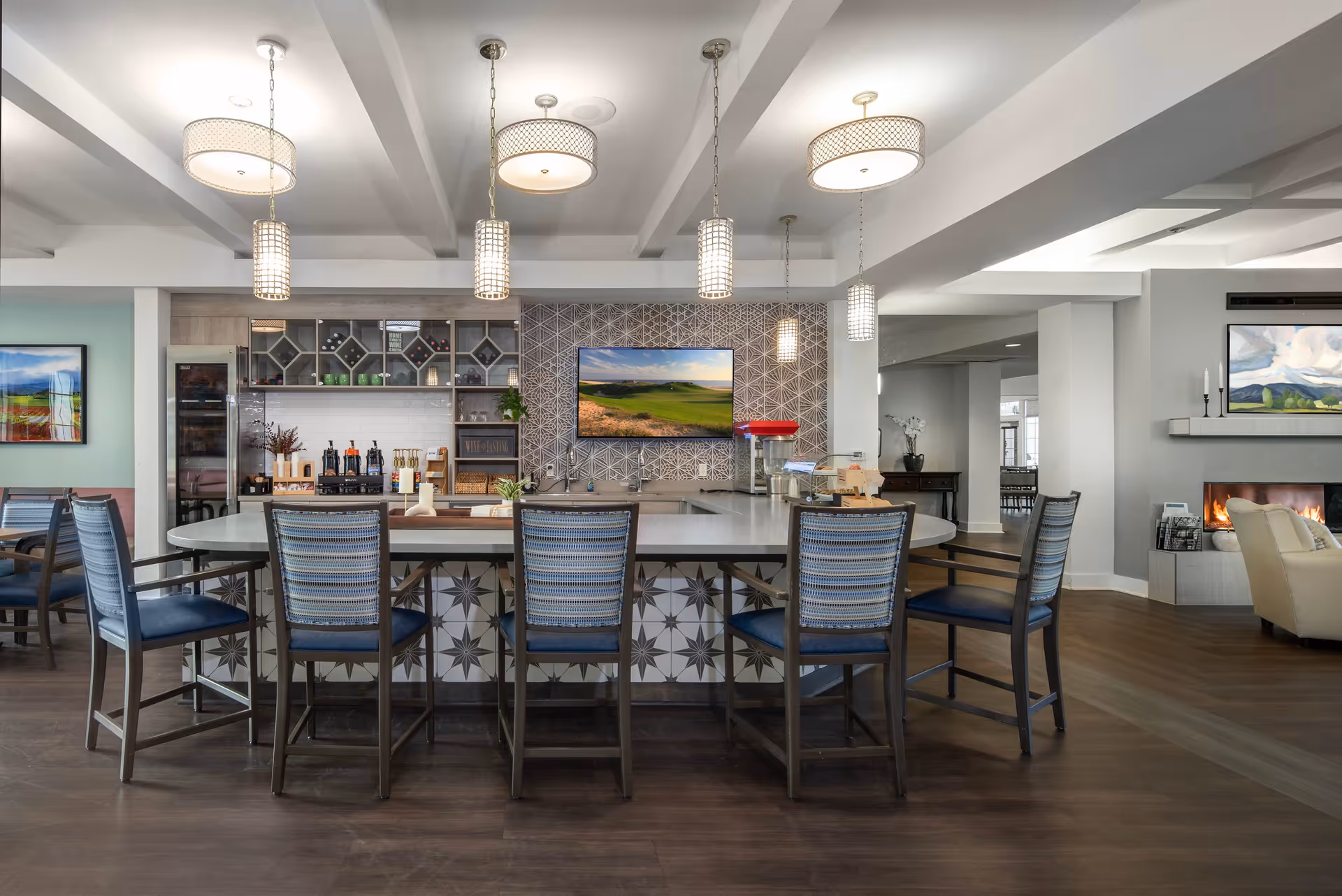 A modern communal dining and lounge area featuring a large oval table with six chairs, pendant lights hanging from the ceiling, a wall-mounted TV, a fireplace, and decorative artwork on the walls. The space has a warm and inviting atmosphere with wood flooring and patterned tiles on the table base.