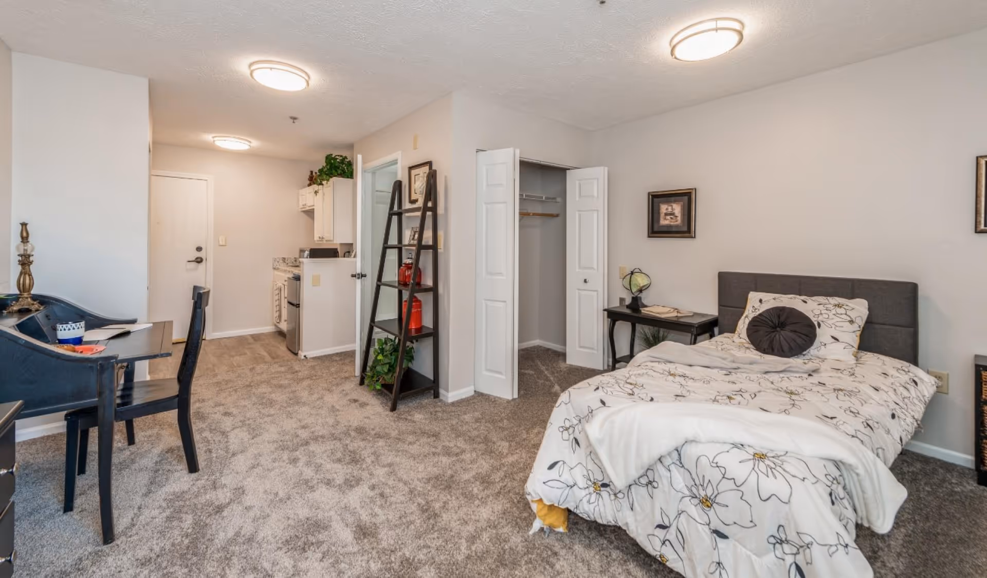 A cozy studio apartment featuring a bed with floral bedding, a small bedside table with a lamp, an open closet with double doors, a black ladder-style shelf with decorative items, a desk with a chair, and a kitchenette in the background. The room has beige walls and carpeted flooring.