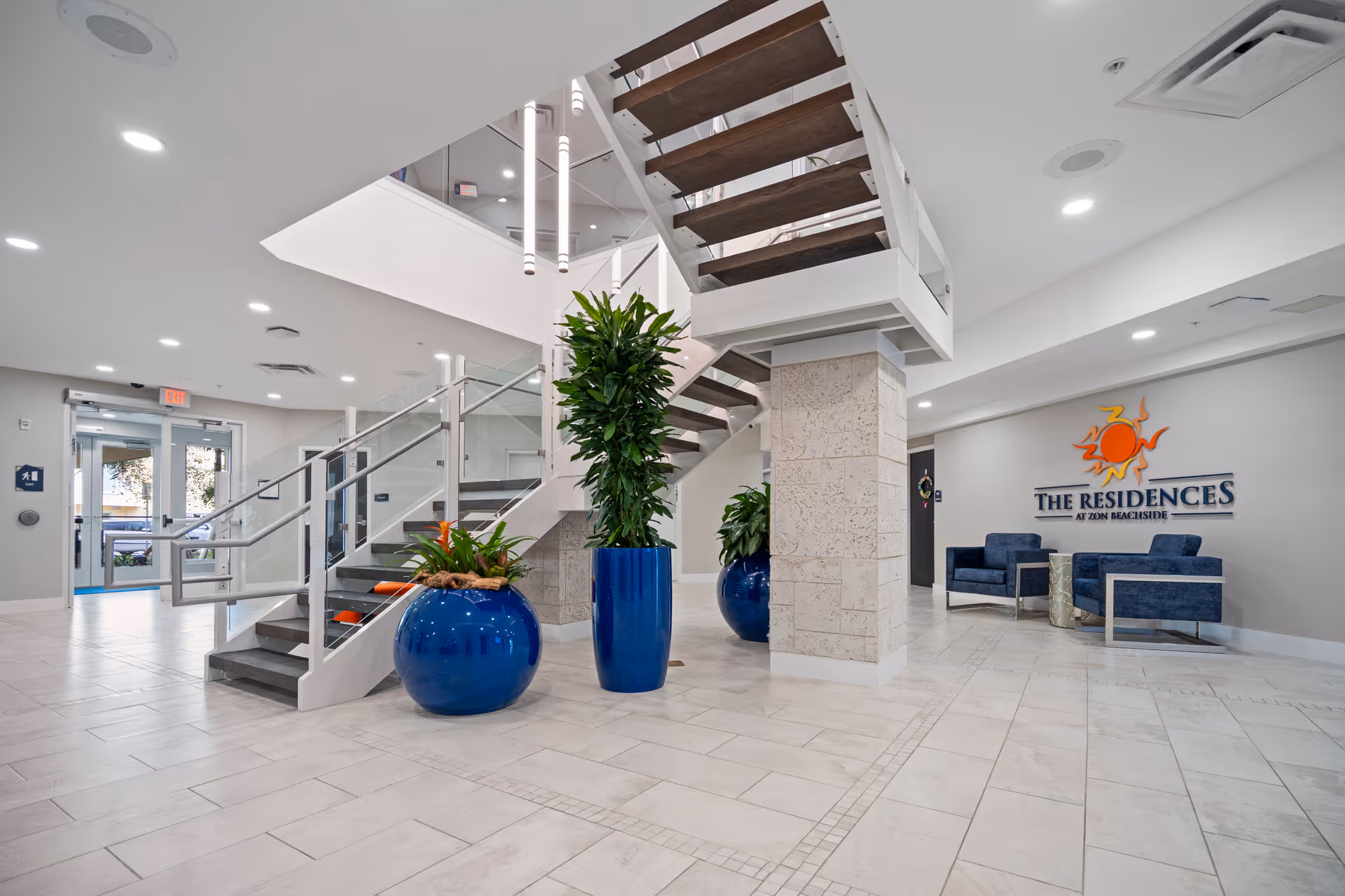 Bright and modern lobby area of The Residences at Zon Beachside featuring a staircase with glass railings, large blue planters with green plants, tiled floor, and a seating area with blue chairs near a wall sign displaying the facility's name and logo.