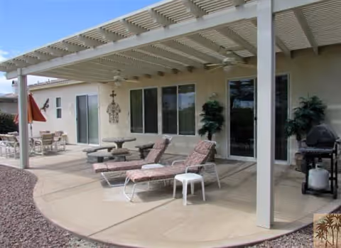 Outdoor patio area with a pergola providing shade over two cushioned lounge chairs and a small white side table. There is a round table with chairs in the background, potted plants near the building wall, and a barbecue grill on the right side. The patio is adjacent to a beige building with multiple windows and a sliding glass door.