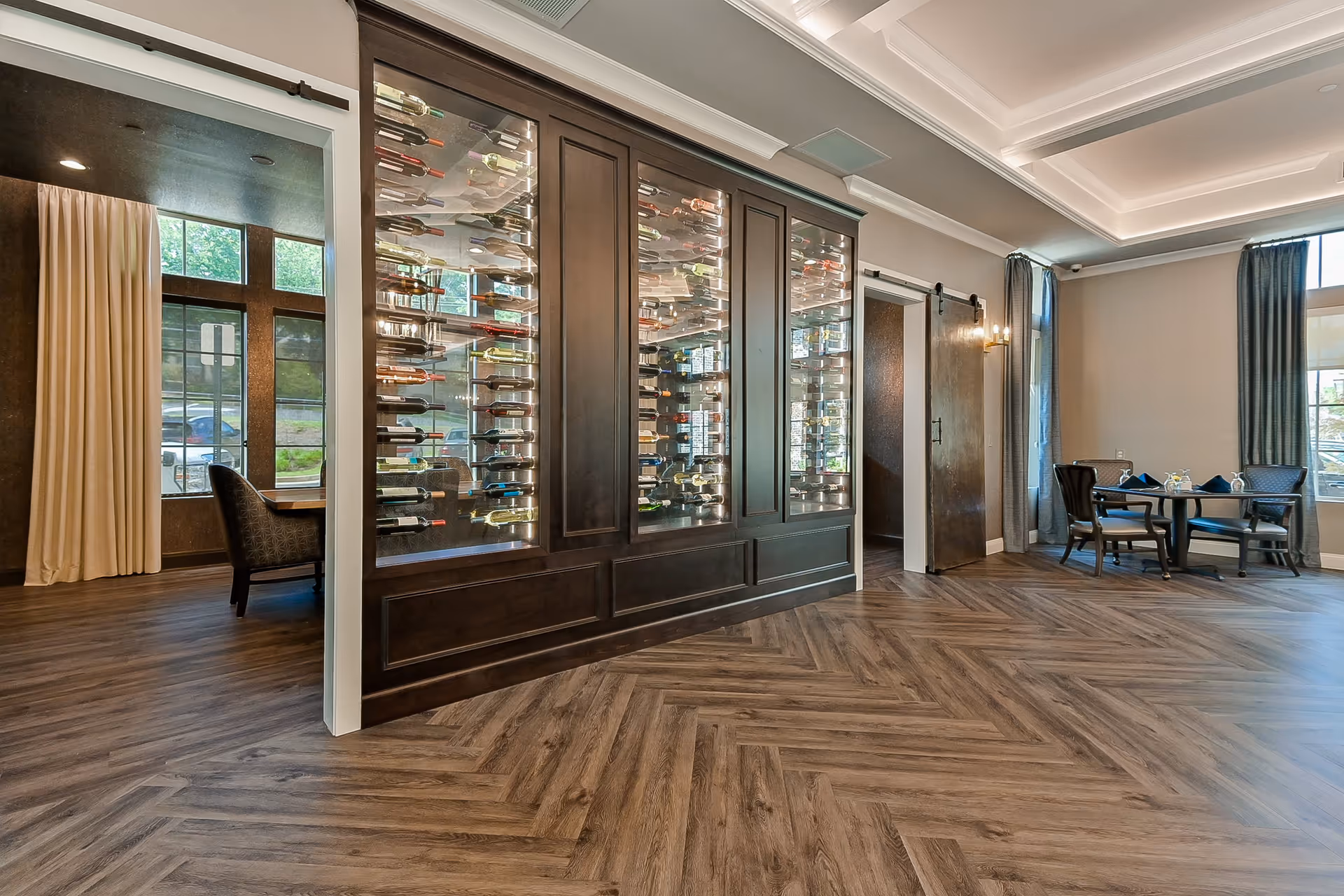 Interior of a senior living facility featuring a modern wine display cabinet filled with bottles, a small dining table with four chairs near large windows with curtains, and a room with an armchair visible through an open doorway. The space has wood-patterned flooring and a coffered ceiling with recessed lighting.