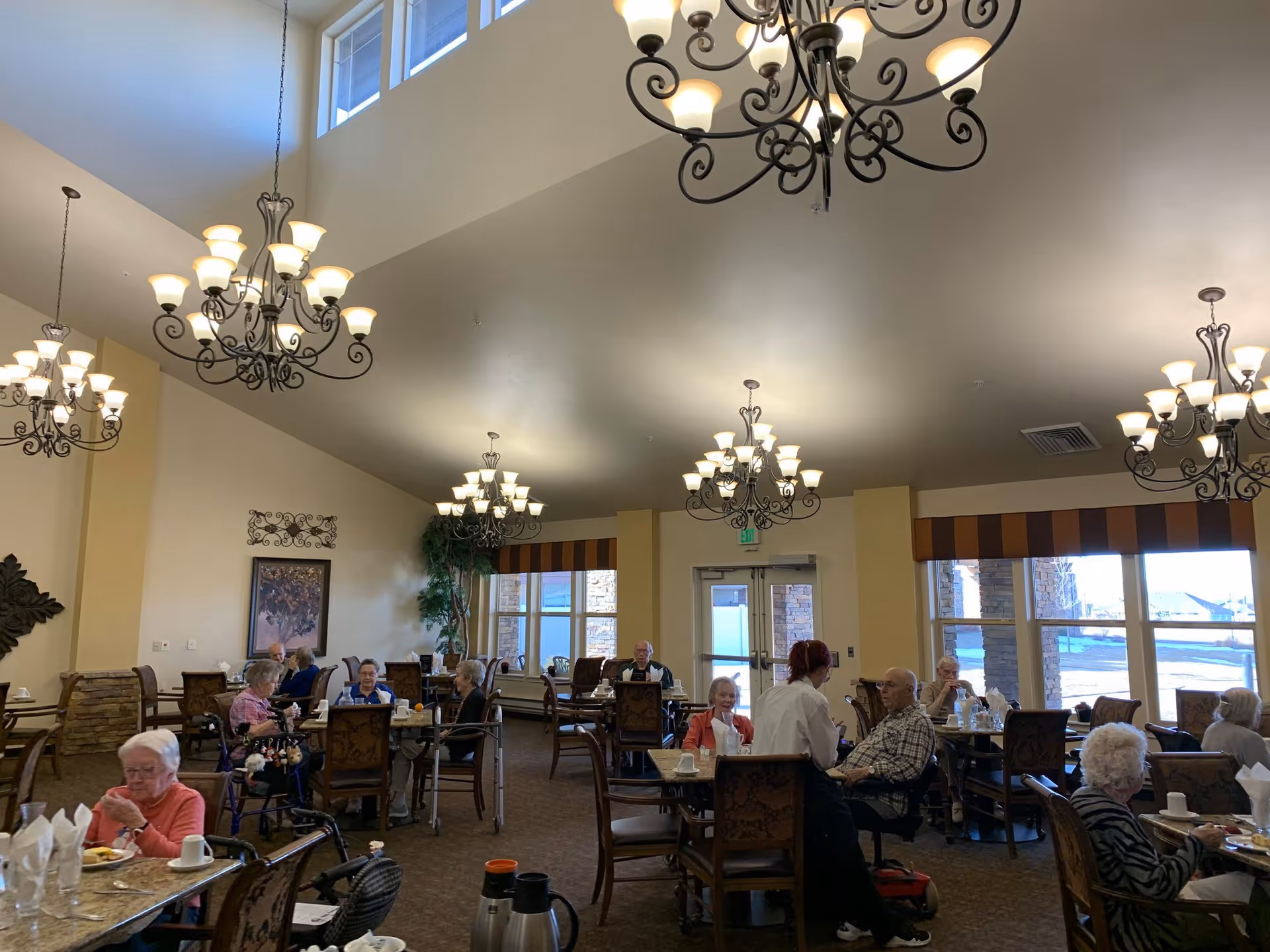 A dining room in a senior living facility with several elderly residents seated at tables, eating and conversing. The room has high ceilings with multiple ornate chandeliers, large windows with striped valances, and warm yellow walls. A staff member is attending to one of the residents.