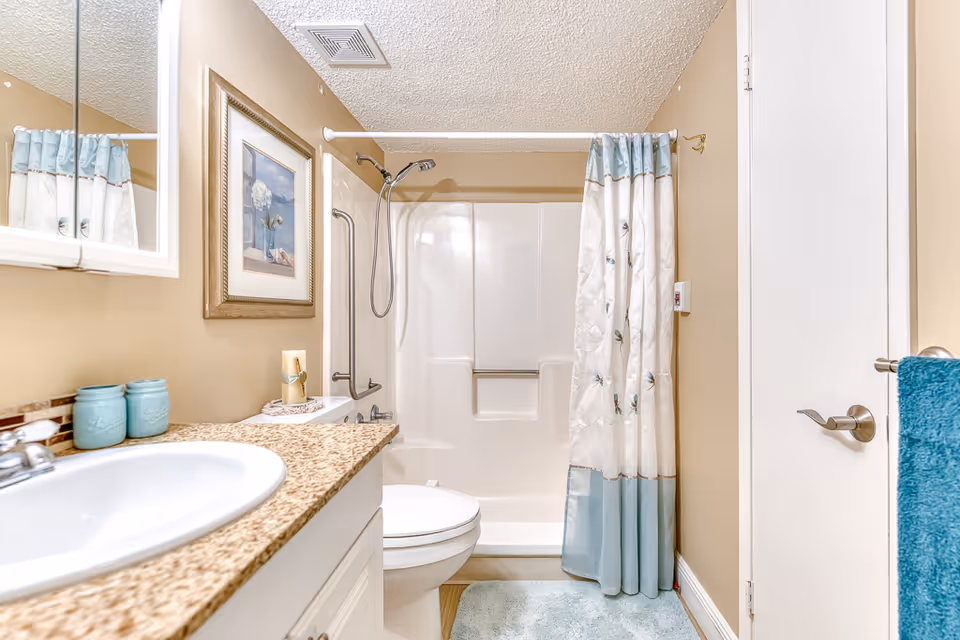 A clean and well-lit bathroom featuring a white sink with a granite countertop, a toilet, and a bathtub with a showerhead and a white and blue floral shower curtain. The walls are painted beige, and there is a framed picture hanging above the toilet. A blue towel hangs on a silver towel rack attached to a white door, and a light blue bath mat is on the floor.