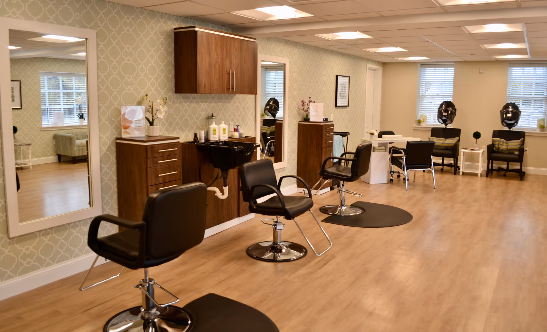 Interior view of a senior living facility's salon area with three black salon chairs in front of mirrors and a sink, wooden cabinets, and hair drying stations near windows. The room has light wood flooring and patterned wallpaper.