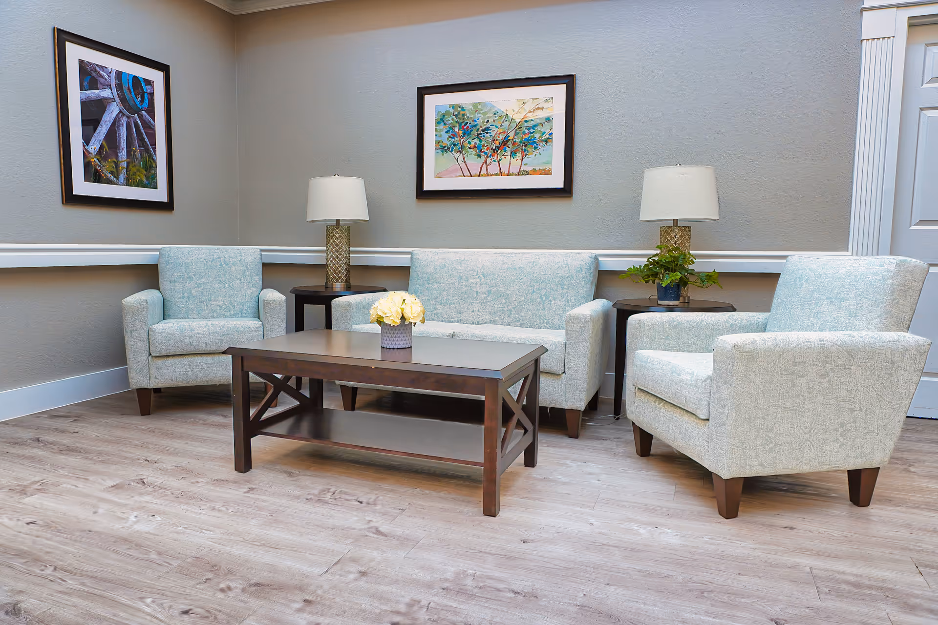 A cozy sitting area with a light blue loveseat and two matching armchairs arranged around a dark wooden coffee table with a small flower vase. Two side tables with lamps and a small plant are placed beside the loveseat and armchairs. The walls are painted gray and decorated with two framed pictures, one of a wagon wheel and the other of a colorful tree landscape.