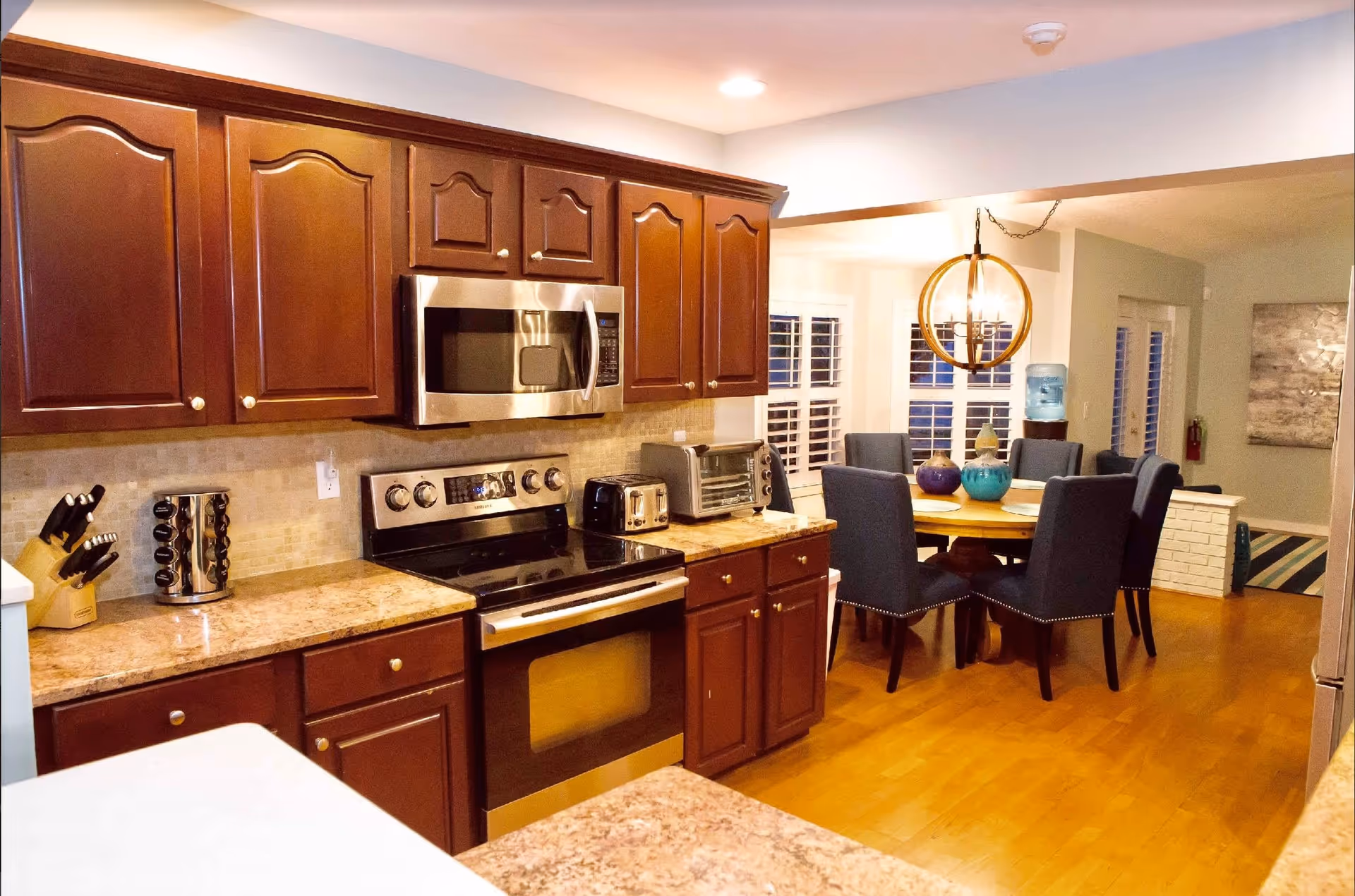 Interior view of a kitchen with wooden cabinets, a stainless steel microwave and oven, a toaster, a knife block, and a spice rack on the countertop. Adjacent to the kitchen is a dining area with a round wooden table surrounded by six dark upholstered chairs and a modern circular chandelier overhead.