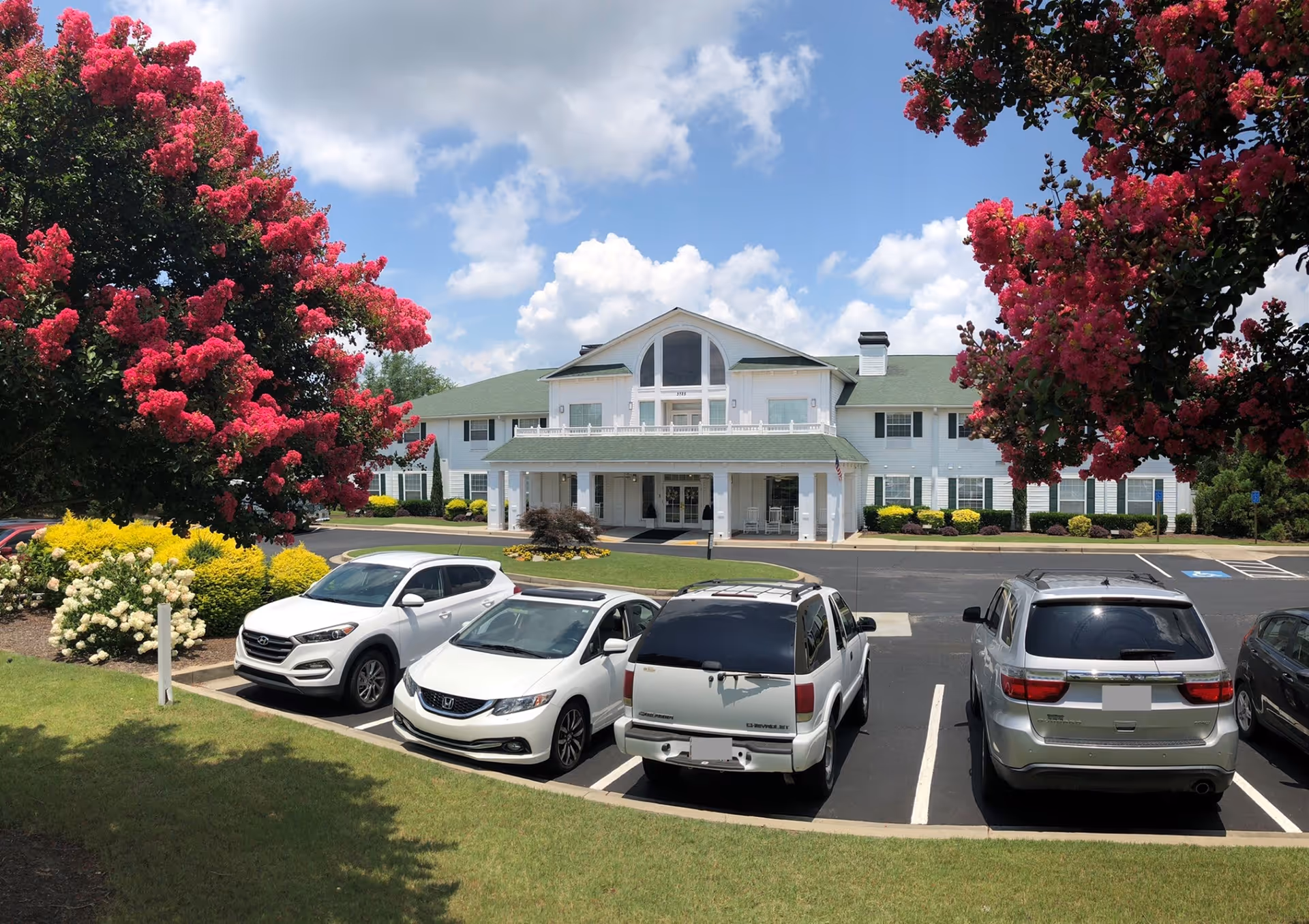 Front view of a two-story white senior living building with a covered entrance, parked cars and blooming pink trees.