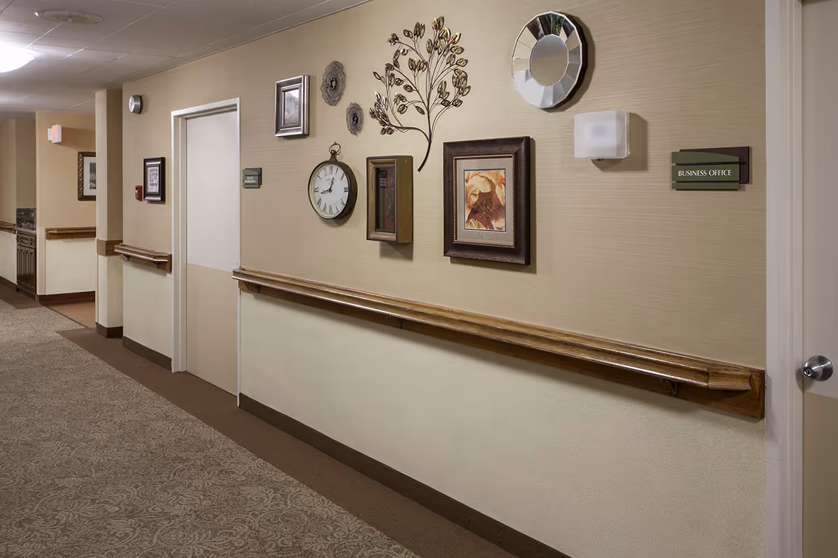 A hallway in a senior living facility with beige walls and carpeted floor. The wall features decorative items including a clock, framed pictures, a metal wall art piece shaped like a tree, and a round mirror. There are wooden handrails along the wall and a door labeled 'Business Office'.