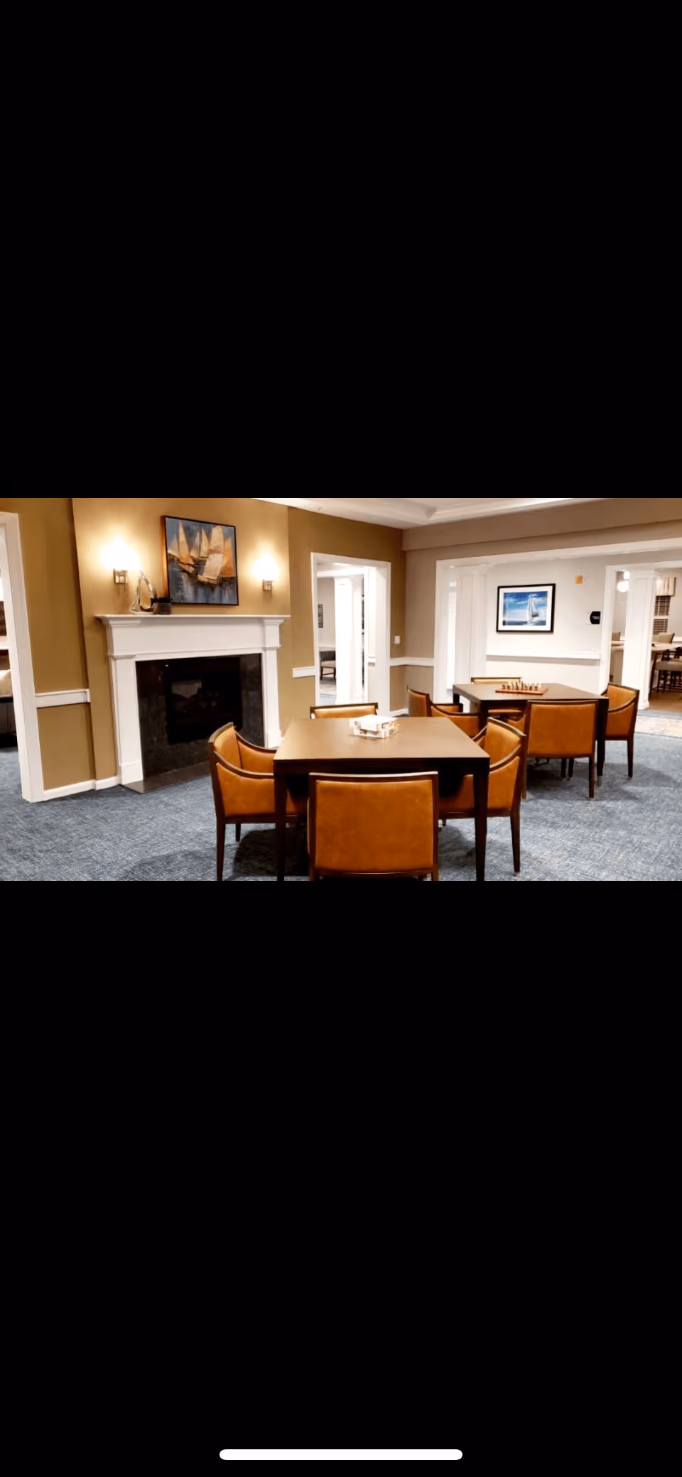 Interior view of a senior living facility common area with multiple square tables surrounded by brown leather chairs. The room features beige walls, a white fireplace with a painting of sailboats above it, and wall sconces on either side of the fireplace. The carpet is blue, and there are doorways leading to other rooms with additional seating and artwork on the walls.