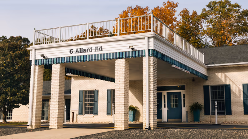 Front entrance of a light-brick building with a covered portico, teal doors and shutters, and a sign reading "6 Allard Rd."