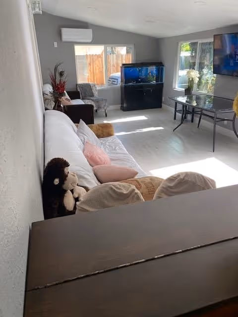 A bright living room with a white couch adorned with pillows and stuffed animals, a glass table with a vase of white flowers, a large aquarium on a black stand, a wall-mounted TV, and two windows letting in natural light.