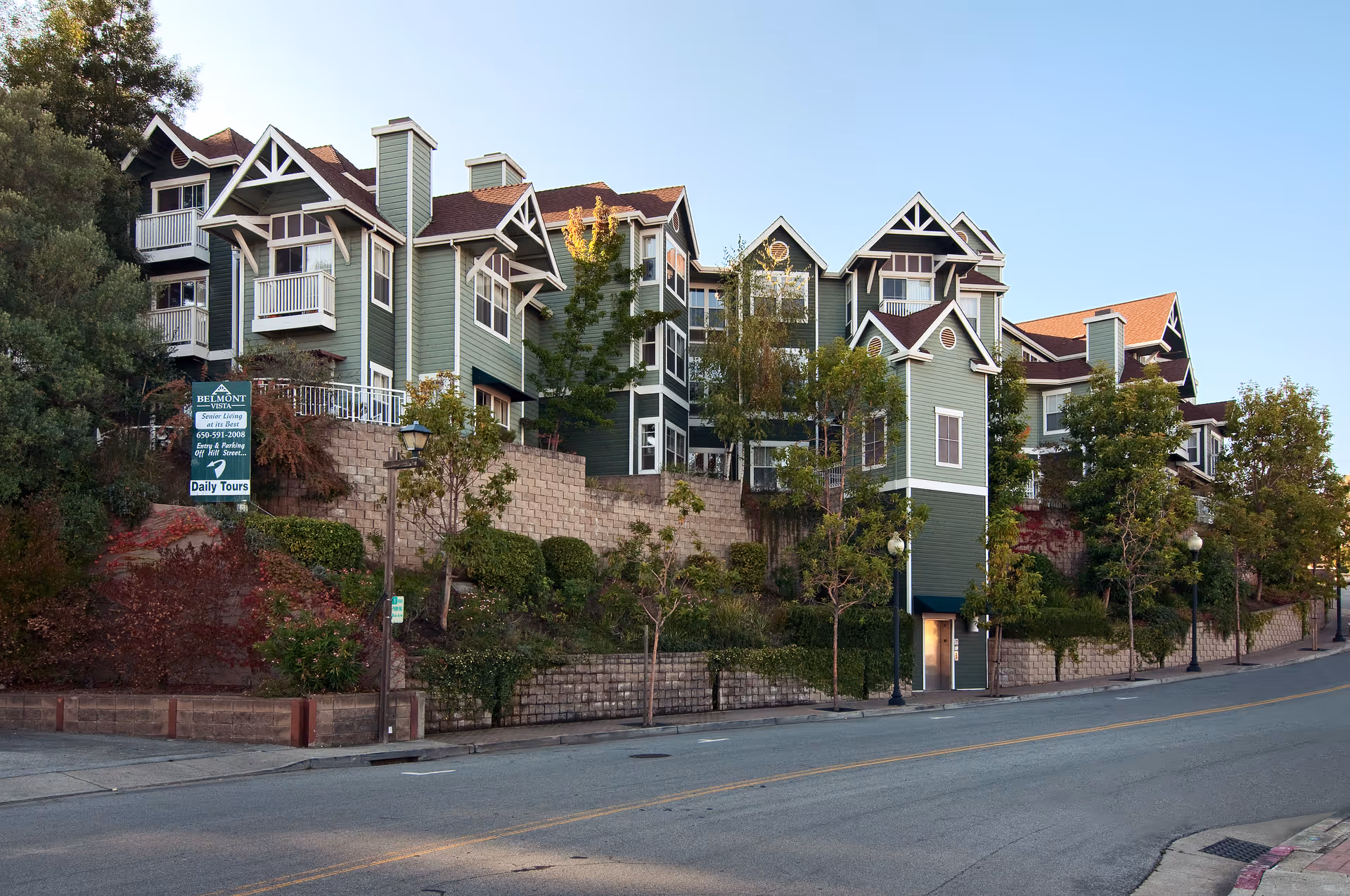Exterior view of a multi-story senior living facility with green siding, white trim, and multiple balconies. The building is situated on a raised landscaped area with trees and shrubs, bordered by a stone retaining wall along a street. A sign near the building advertises Belmont Vista Senior Living with contact information and daily tours.