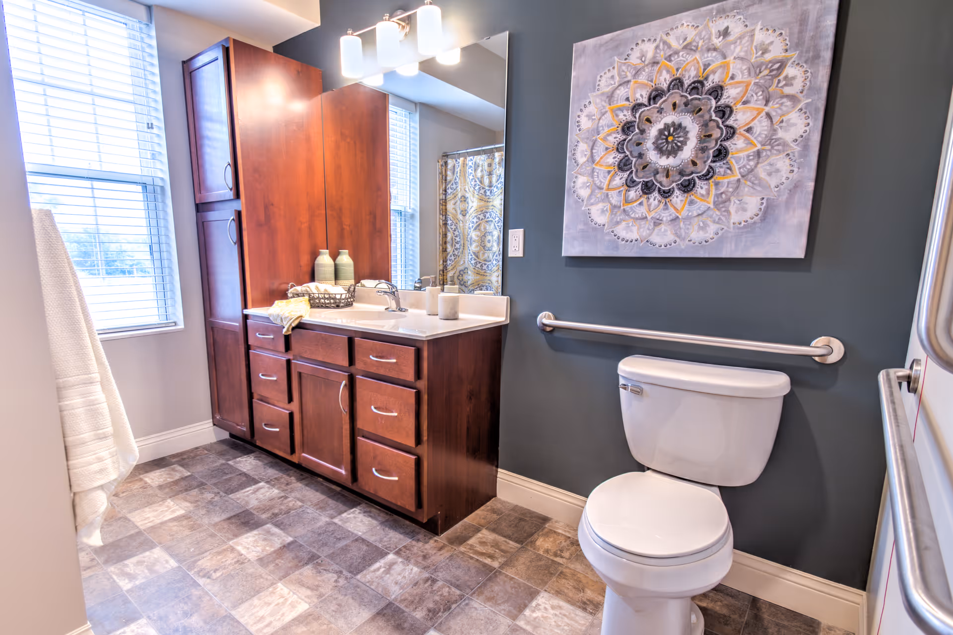A bright bathroom featuring a white toilet with a safety grab bar on the wall behind it, a wooden vanity with multiple drawers and a sink, a large mirror above the sink, a window with blinds, a towel hanging on a rack, and a decorative mandala artwork on the dark-colored wall.