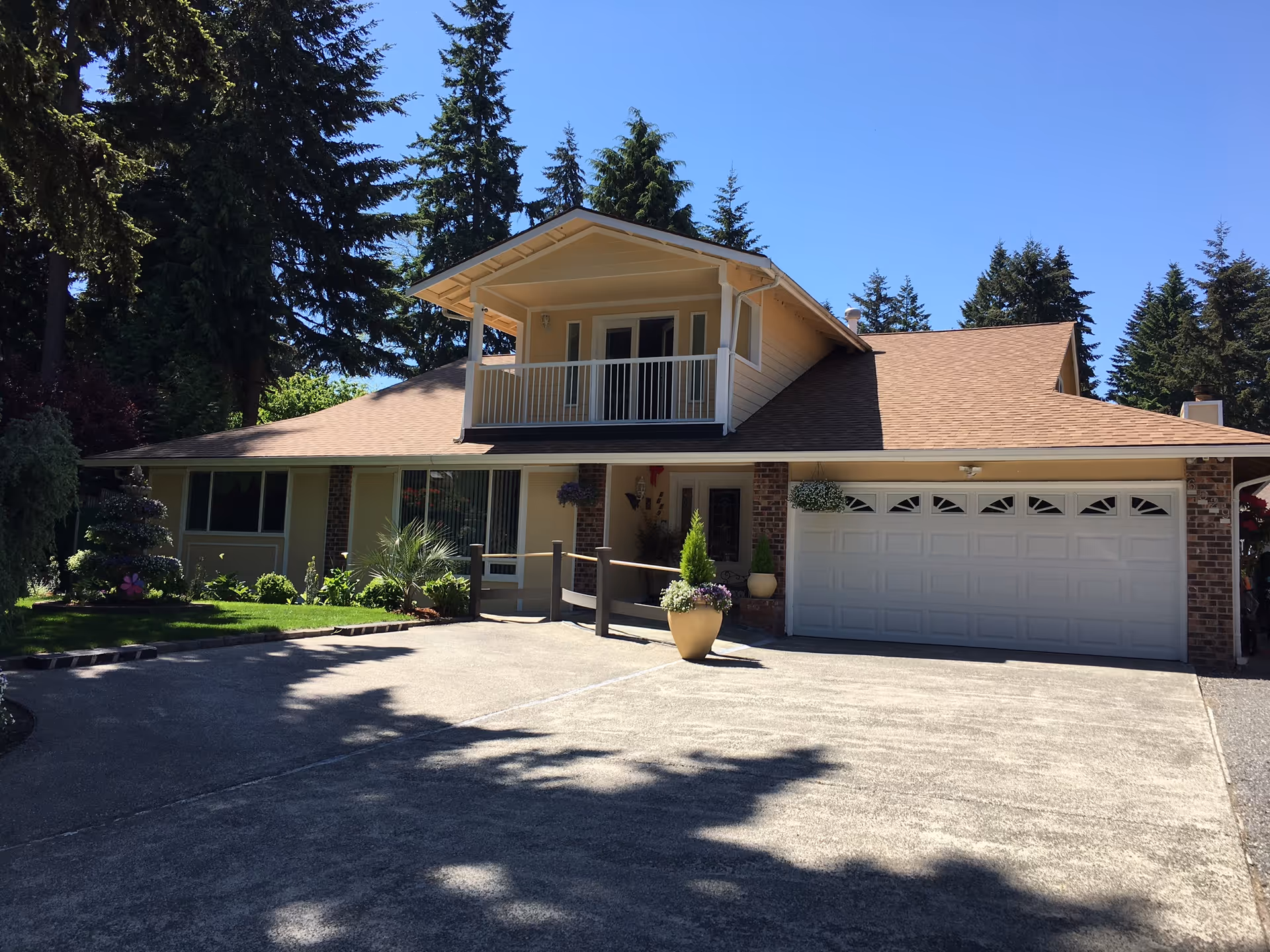A two-story residential building with a beige exterior and brown roof, surrounded by tall pine trees. The house features a balcony on the upper floor, a large garage door on the right, and a driveway in front. There are potted plants and greenery near the entrance, and the sky is clear and blue.