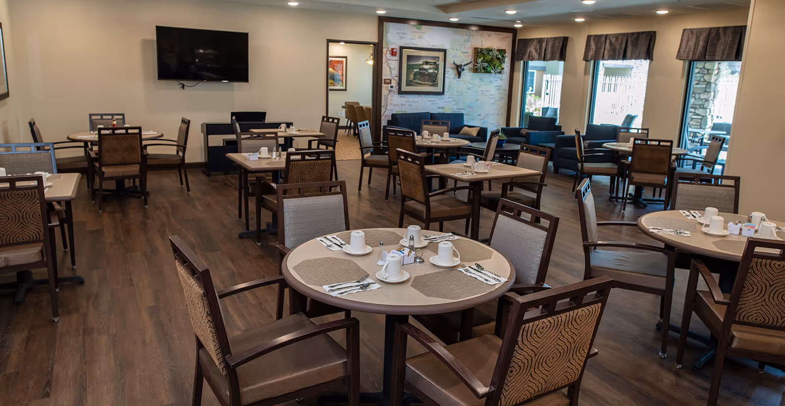 A dining room with multiple round tables set with cups, napkins, and utensils. The room has wooden flooring, several chairs around each table, a wall-mounted TV, and large windows with curtains letting in natural light. There is a seating area with blue sofas and framed artwork on the walls.