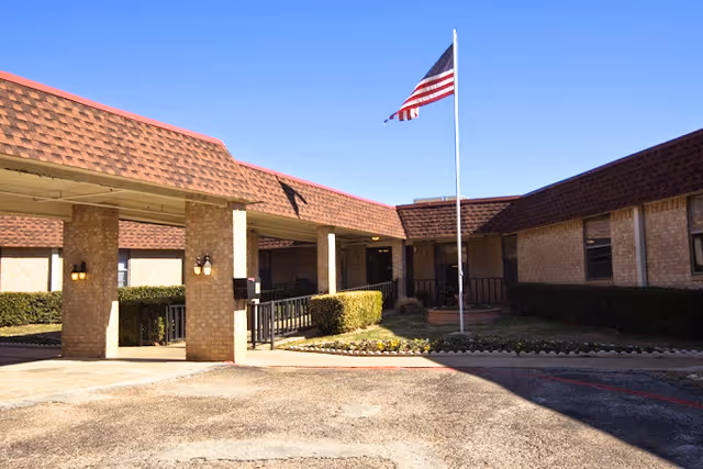 Exterior view of a single-story brick building with a covered entrance supported by columns, an American flag on a flagpole in front, and neatly trimmed bushes along the building under a clear blue sky.