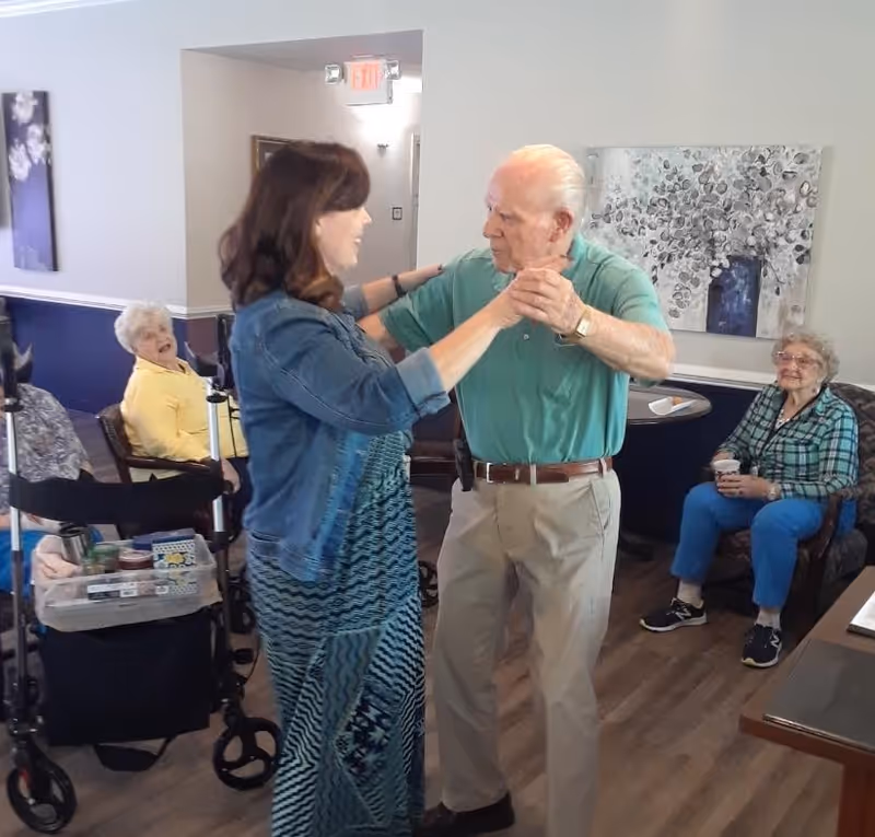 An elderly man and a woman are dancing together in a common area of a senior living facility. Two elderly women are seated nearby, one in a wheelchair and the other on a chair, watching and smiling. The room has wooden flooring, a painting on the wall, and an exit sign above a doorway.