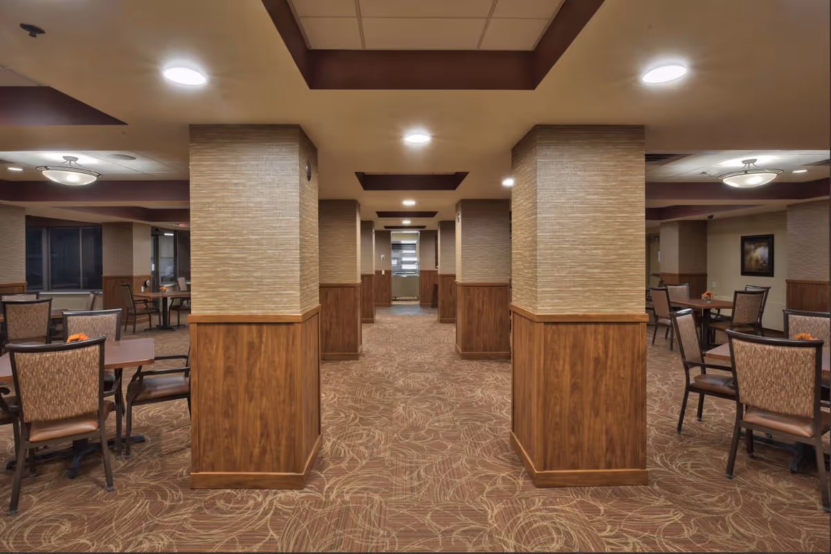 Interior view of a dining area in a senior living facility with multiple tables and chairs arranged around square tables. The room features carpeted flooring with a leaf pattern, beige walls with wooden paneling on the lower half, and several large square columns. Ceiling lights provide illumination, and there is a framed picture on one wall.