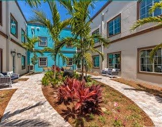 Outdoor courtyard area of a senior living facility with a paved walkway, tropical plants including palm trees and red foliage, surrounded by a two-story building with windows and outdoor seating.
