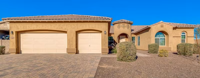 Front exterior of a single-story tan stucco house with a three-car garage and desert landscaping under a clear blue sky.