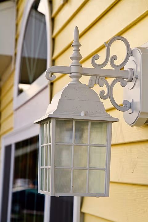 Close-up of a white decorative outdoor wall lantern mounted on a yellow wooden exterior wall near a window.