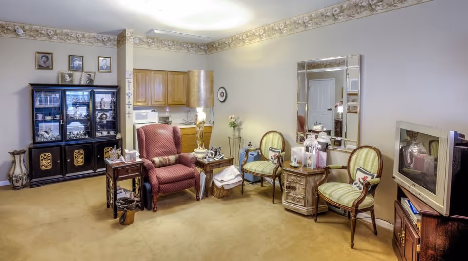 A cozy living room area with a red upholstered armchair, two green striped chairs, a wooden cabinet with glass doors displaying decorative items, a large mirror on the wall, a TV on a wooden stand, and a small kitchenette with wooden cabinets in the background. The room has beige carpet and floral wallpaper border near the ceiling.