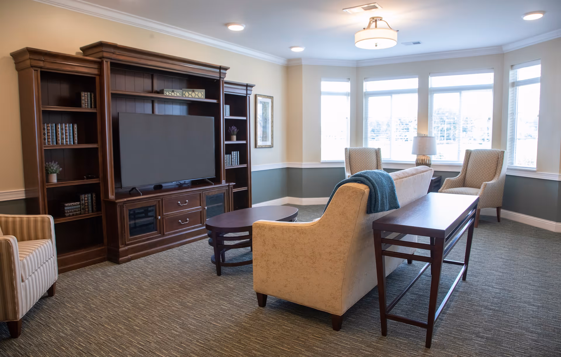 A well-lit living room with a large wooden entertainment center holding a flat-screen TV and decorative books. The room features a beige sofa with a blue throw blanket, two beige armchairs near a bay window, a wooden coffee table, and a side table with a lamp. The walls are painted in a two-tone style with cream on top and green on the bottom, and the floor is carpeted.