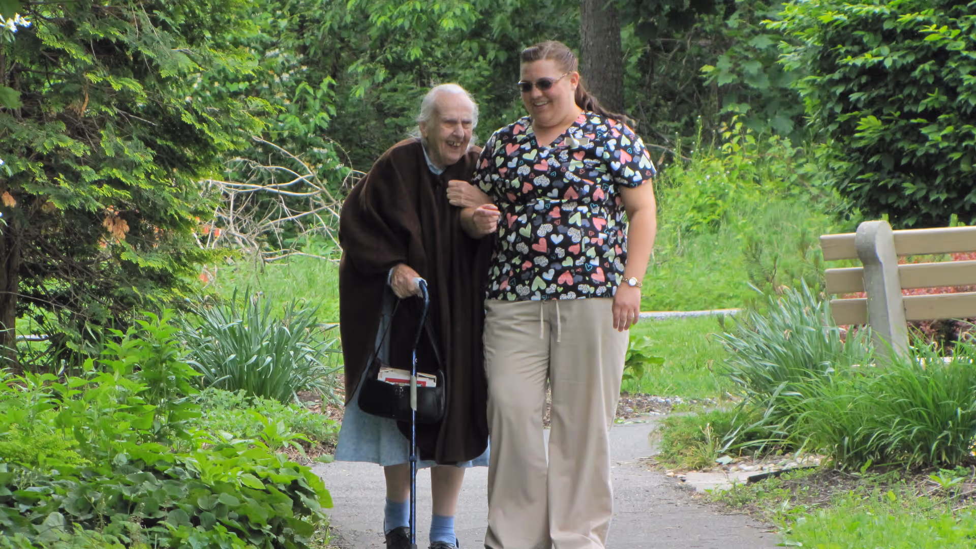An elderly woman using a cane is walking outdoors on a paved path, supported by a caregiver wearing a colorful heart-patterned scrub top and beige pants. They are surrounded by lush green plants and trees, with a wooden bench visible on the right side.