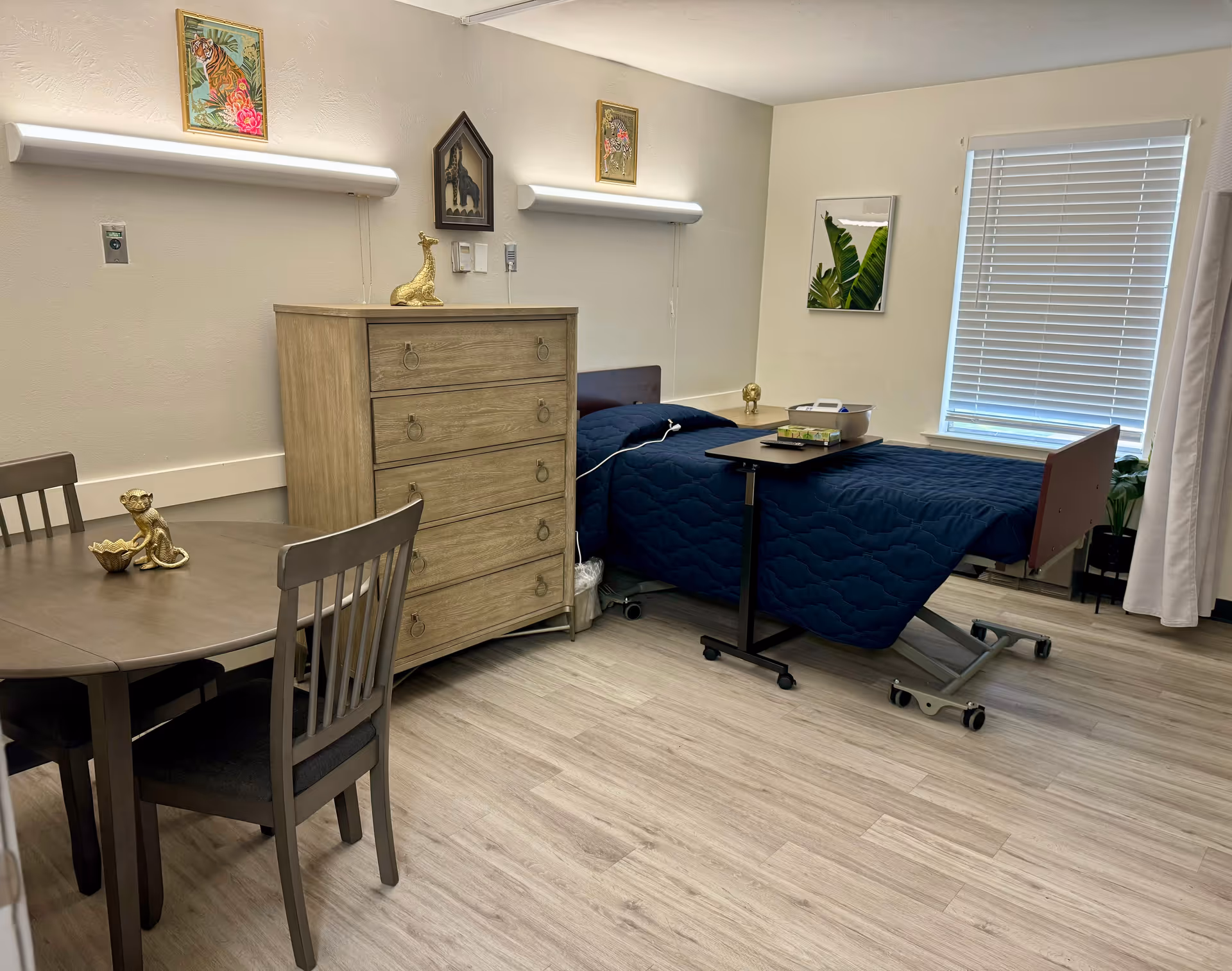 A senior living facility bedroom with a single bed covered in a dark blue quilt, a small overbed table with books and a box, a wooden dresser, a round wooden table with two chairs, and decorative items including small animal figurines and framed artwork on the walls. A window with white blinds is on the right side, and the floor is light-colored wood.