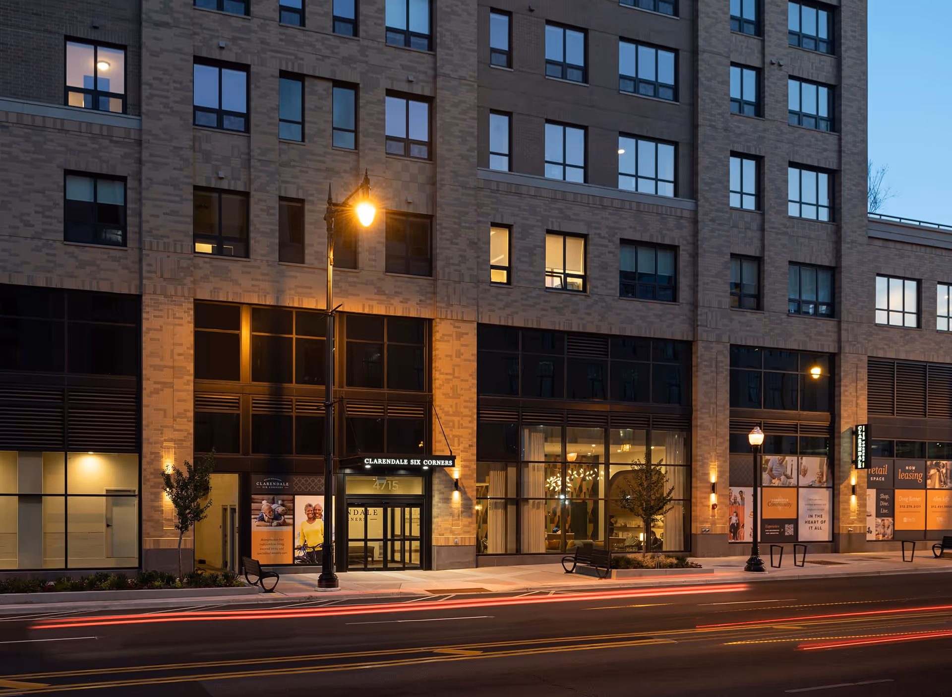 Exterior view of Clarendale Six Corners building at dusk with illuminated entrance and large windows showing interior lighting. Street lamps and benches are visible on the sidewalk in front of the building.