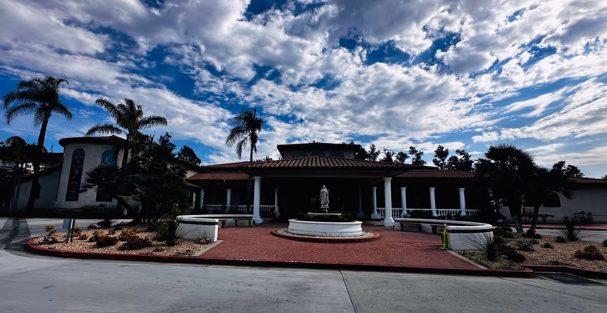 Front entrance of a retirement home with a circular fountain and statue, red brick driveway, and palm trees under a cloudy sky.