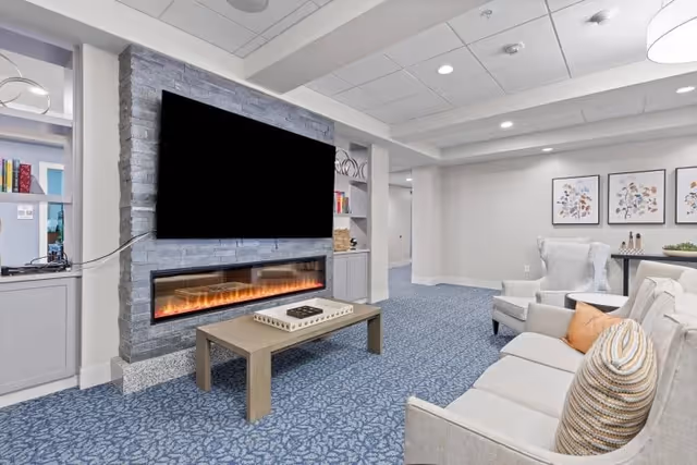 Bright communal living room with a sofa and armchairs facing a wall-mounted TV above an electric fireplace and a central coffee table.