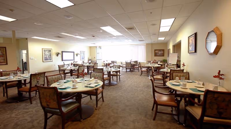 A spacious dining room in a senior living facility with multiple round tables set with plates, cups, and utensils. The room has carpeted floors, beige walls adorned with framed artwork and a mirror, and large windows letting in natural light. Chairs with cushioned seats and backs surround each table.