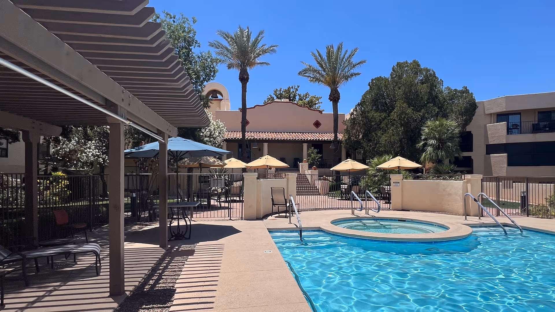 Outdoor swimming pool area at a senior living facility with a hot tub adjacent to the pool. There are several lounge chairs and tables with umbrellas around the pool. Palm trees and other greenery surround the area, with a building featuring a tiled roof and balconies in the background under a clear blue sky.