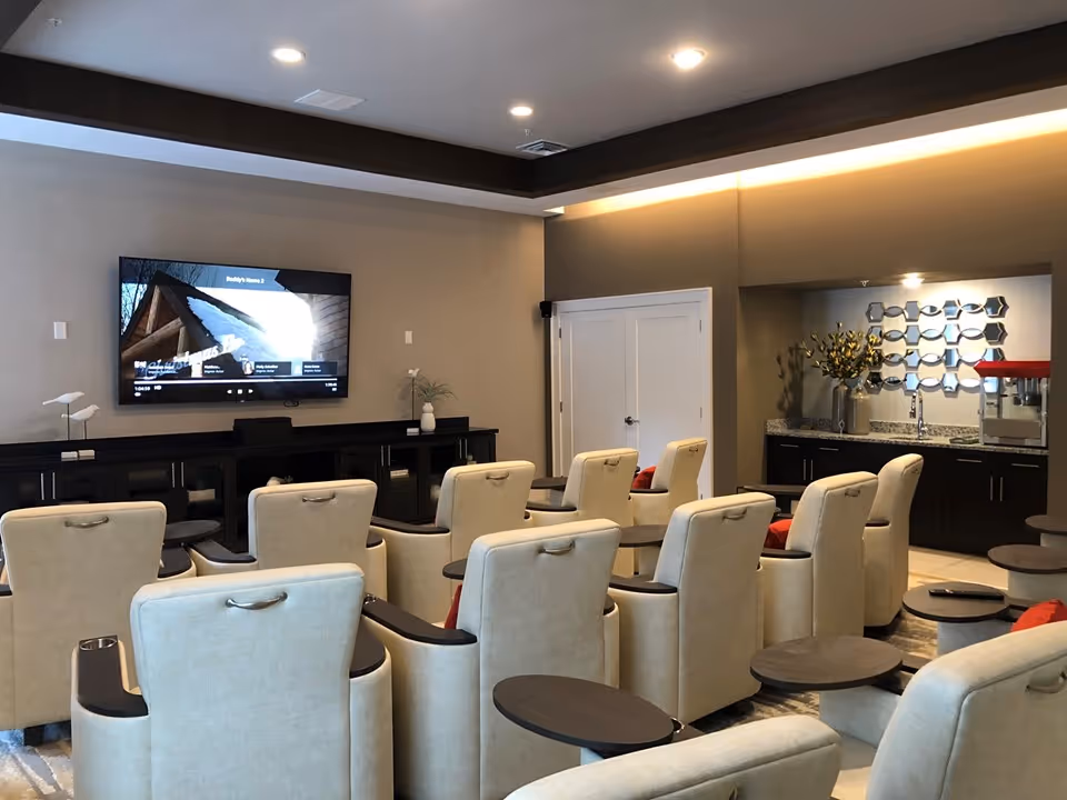 A cozy media room with multiple beige recliner chairs arranged in rows facing a wall-mounted flat-screen TV. The room has a modern design with a dark wood ceiling trim, a side counter with a sink, decorative mirror, and a popcorn machine. The walls are painted in neutral tones, and the lighting is warm and recessed.
