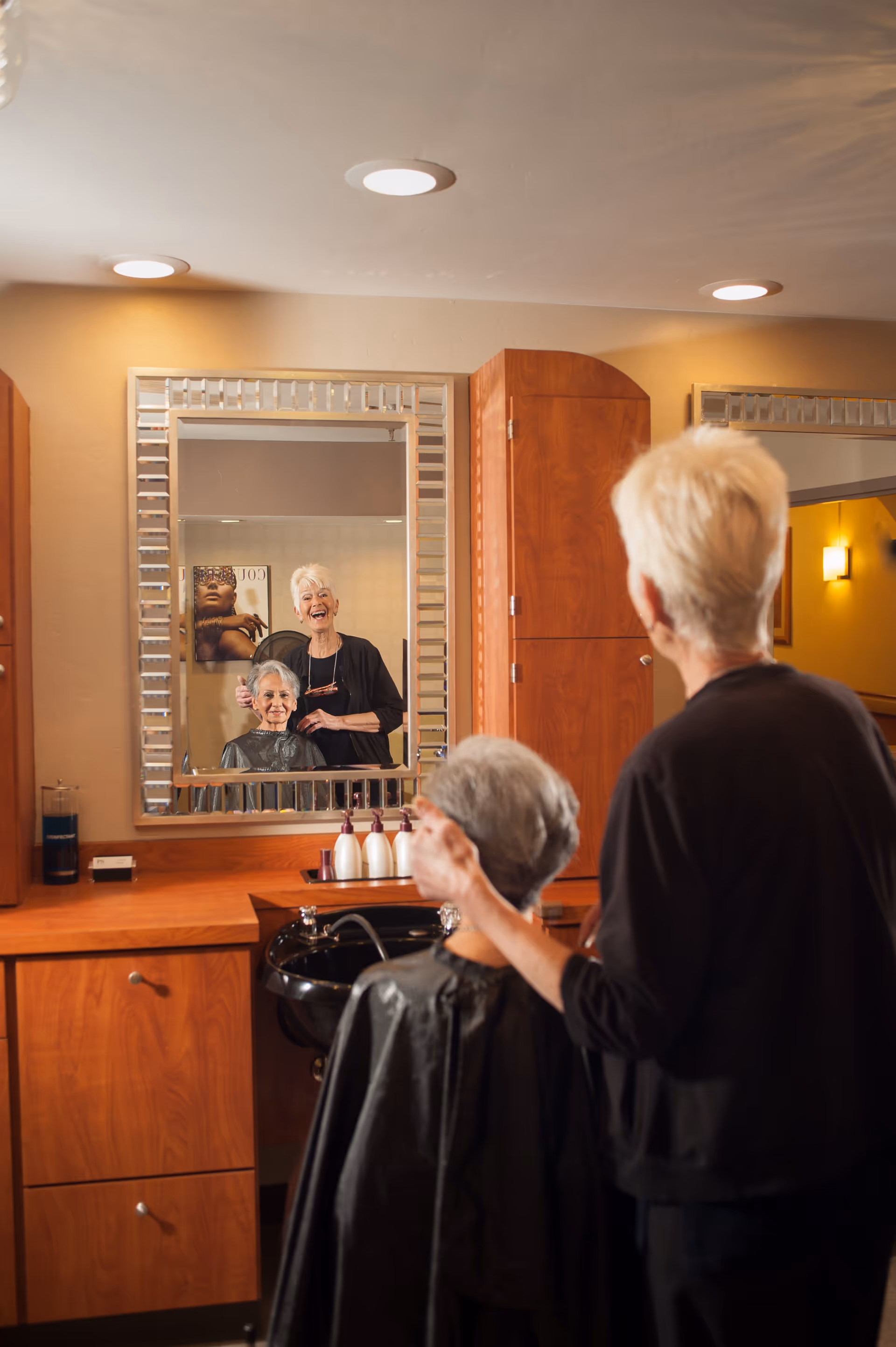An elderly woman is seated in a salon chair with a black cape, getting her hair styled by a hairdresser with short blonde hair. They are in a salon area with wooden cabinets and a large mirror reflecting their smiling faces.