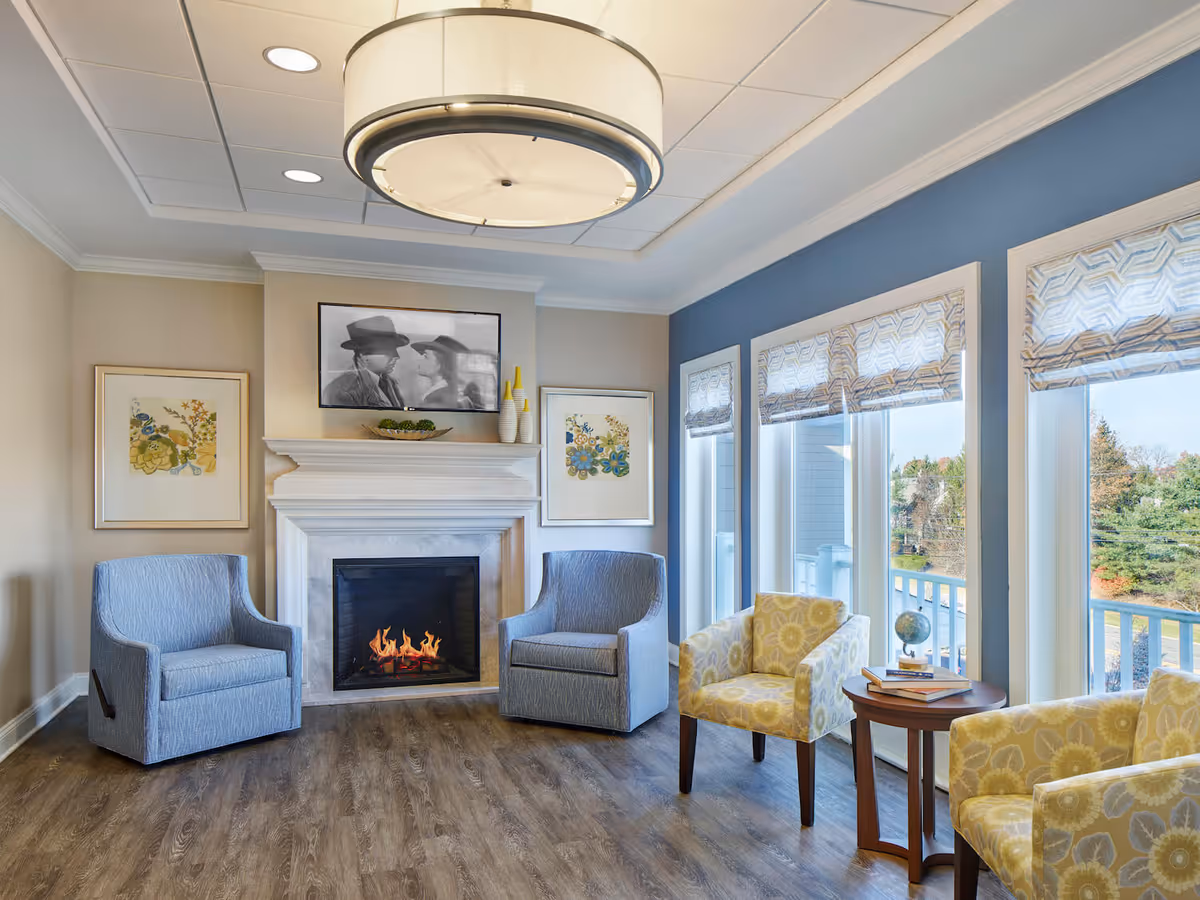 A cozy living room with a lit fireplace, two blue armchairs on either side of the fireplace, and two yellow floral-patterned chairs near large windows with patterned Roman shades. A round wooden side table with books and a small globe is between the yellow chairs. Above the fireplace is a black and white framed photo and decorative vases on the mantel. The room has wood flooring and a modern ceiling light fixture.