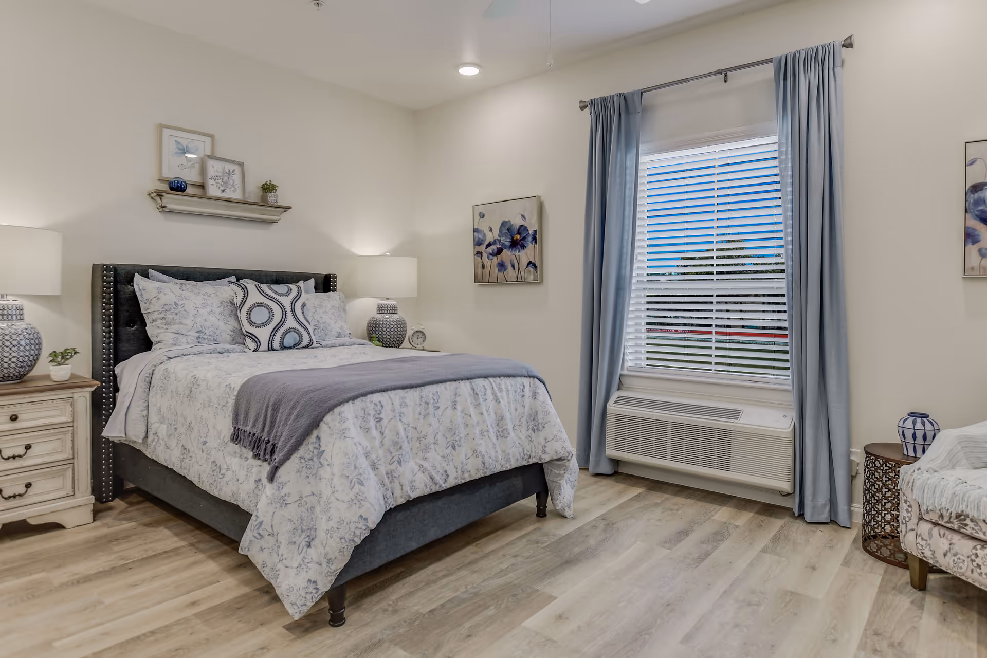 A cozy bedroom with a neatly made bed featuring floral patterned bedding and decorative pillows. The room has light-colored walls, wood flooring, two bedside tables with matching lamps, a window with blue curtains and blinds, and floral artwork on the walls.