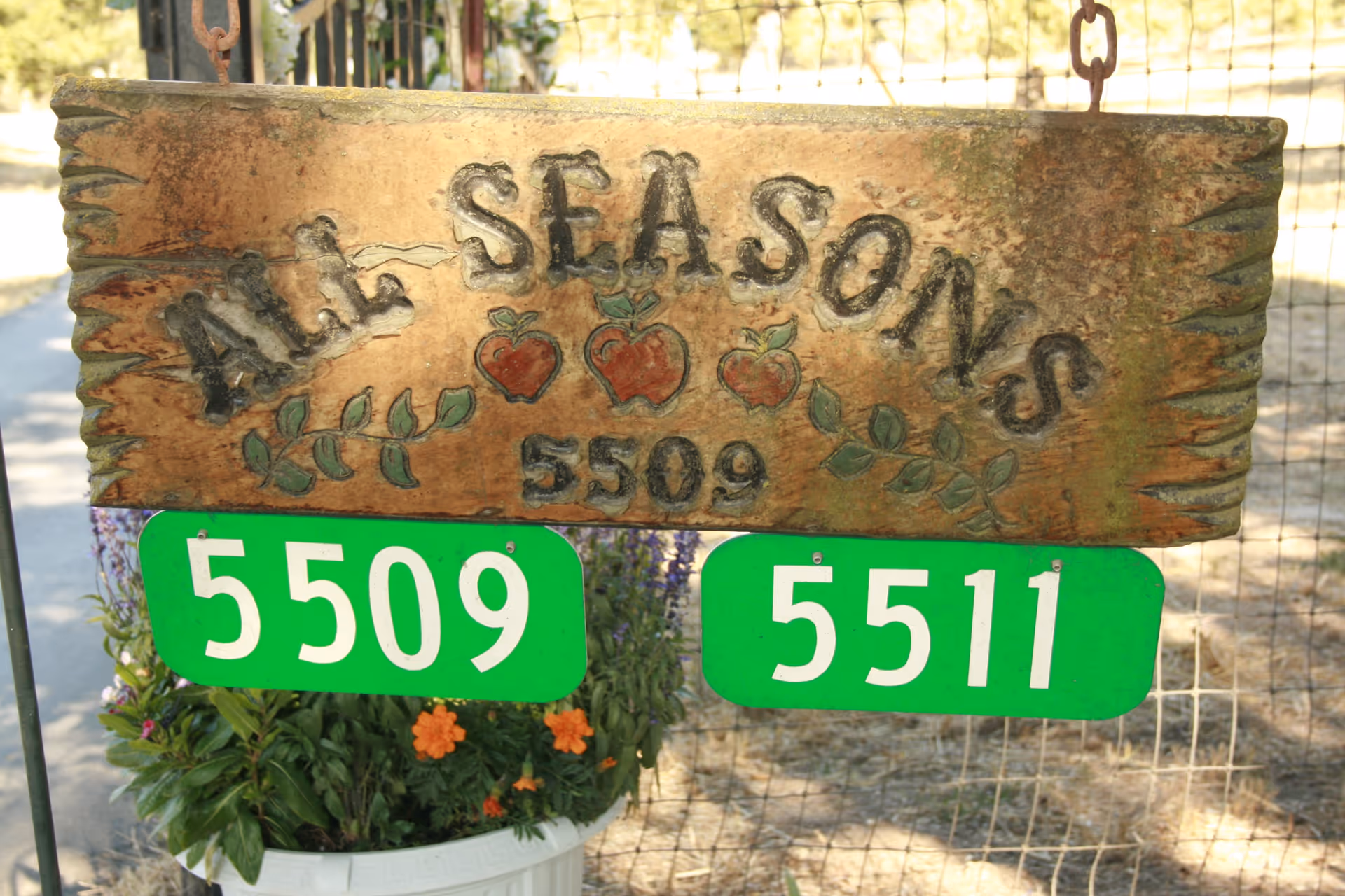 A rustic wooden sign hanging from chains with the text 'ALL SEASONS 5509' and three painted apples with leaves. Below the wooden sign are two green address number signs displaying 5509 and 5511. There are flowers and greenery in a white pot beneath the signs, and a wire fence and outdoor area in the background.