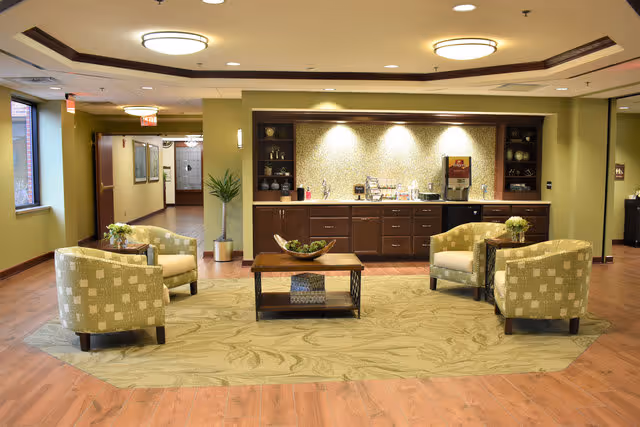 A cozy seating area in a senior living facility with four patterned armchairs arranged around a wooden coffee table on a large area rug. Behind the seating area is a built-in cabinet with a countertop, coffee machine, and various decorative items. The space has warm lighting, wood flooring, and green walls, with a hallway visible in the background.