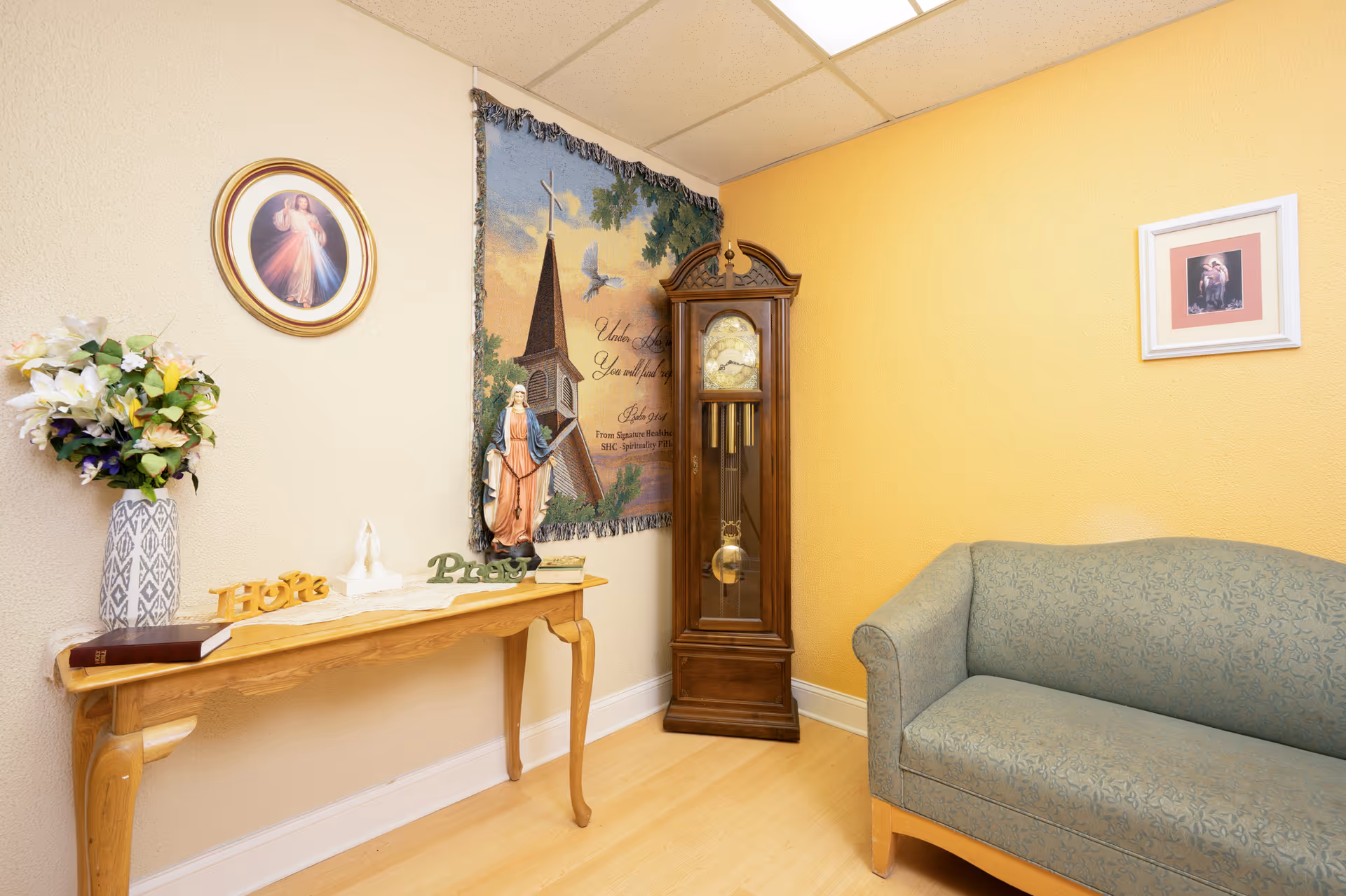 A cozy corner of a room with a wooden grandfather clock, a green patterned couch, and a wooden console table decorated with a vase of flowers, religious figurines, and words 'Hope' and 'Pray'. The walls are painted yellow and beige, adorned with religious artwork and a tapestry featuring a church steeple and a dove.