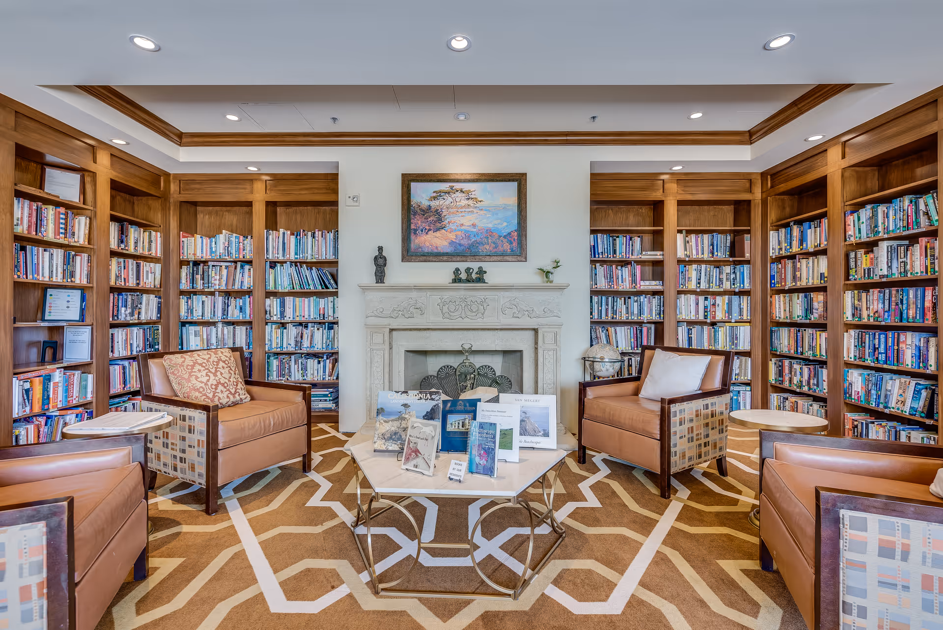 A cozy library room with wooden bookshelves filled with books lining the walls. Four leather armchairs with patterned cushions are arranged around a central coffee table displaying various books and pamphlets. A decorative fireplace with a framed painting above it is centered on the far wall. The room has a patterned carpet and recessed ceiling lights.