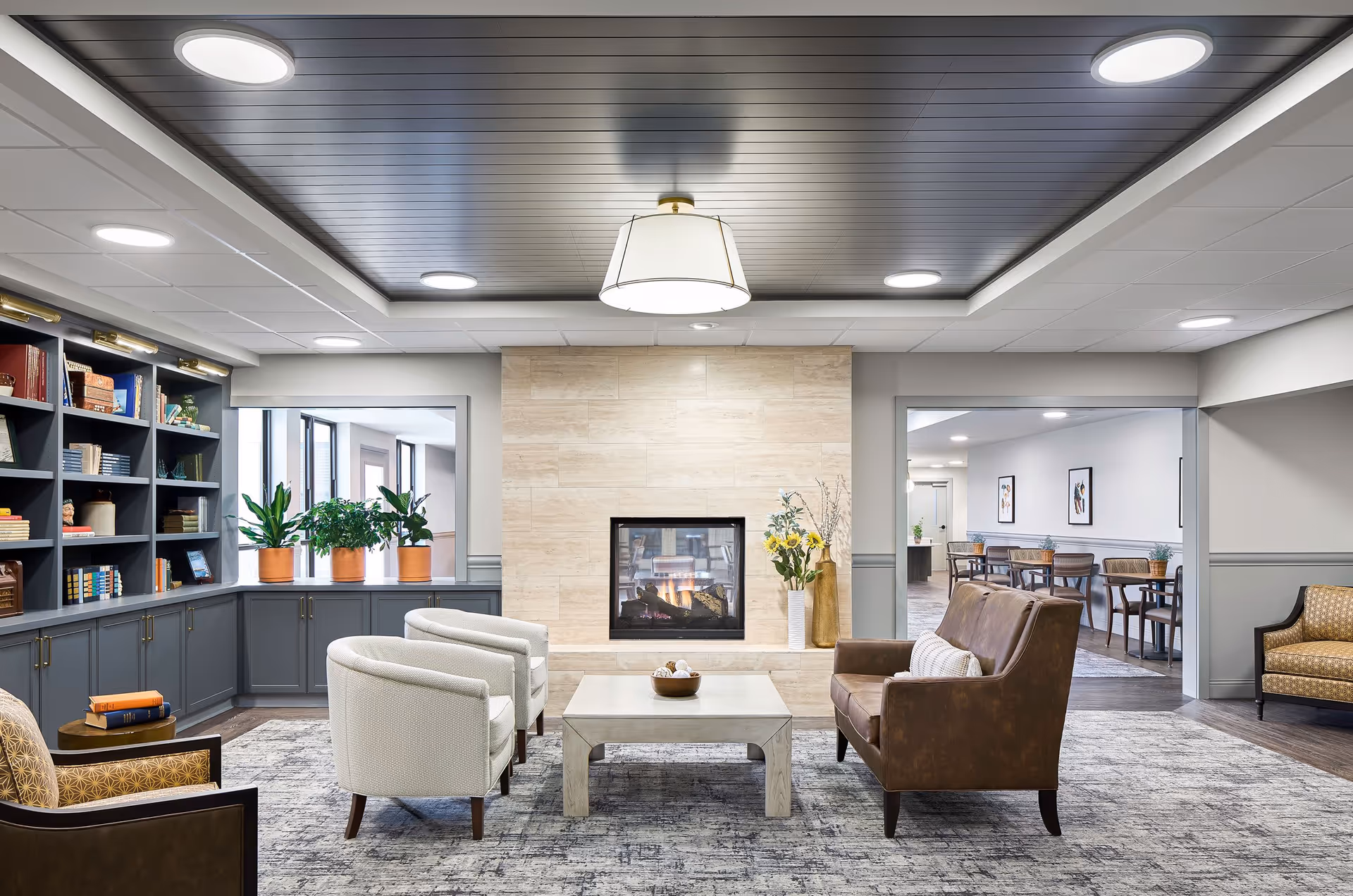 A cozy living room area in a senior living facility featuring a modern fireplace with beige stone surround, a brown leather loveseat, two white armchairs, a wooden coffee table, and built-in dark gray bookshelves filled with books and decorative items. There are potted plants on the windowsill and additional seating visible in an adjoining room.