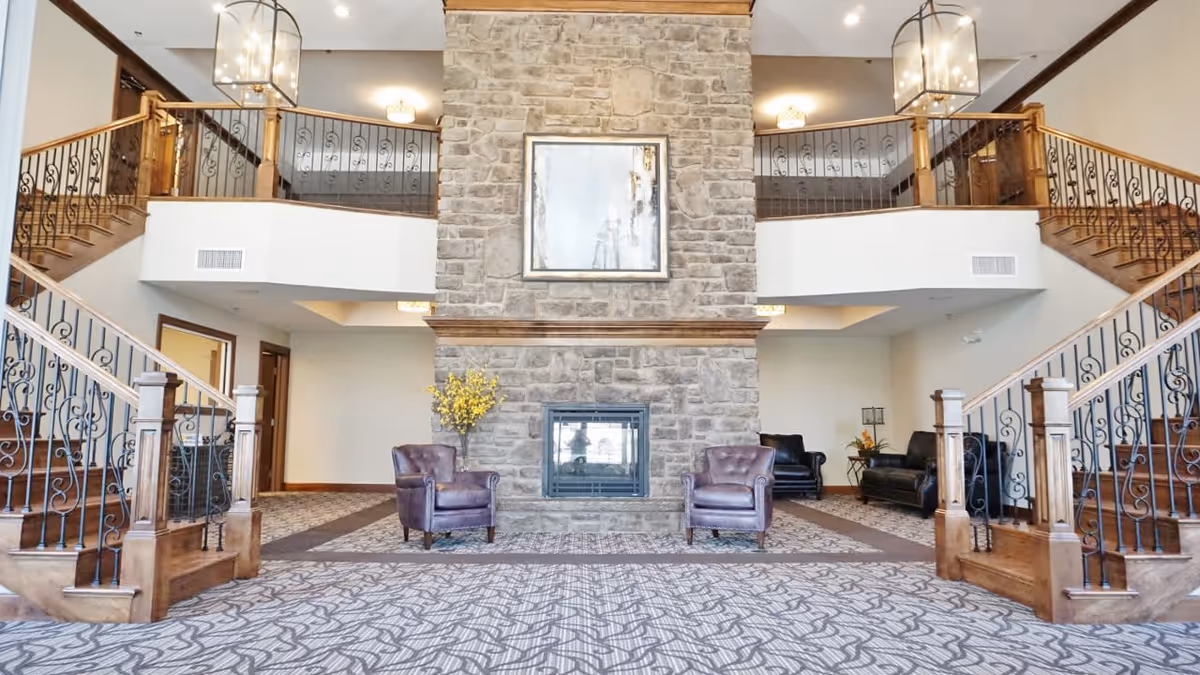 Spacious interior of The Belvedere featuring a large stone fireplace in the center with two purple armchairs in front. The room has a patterned carpet floor, wooden staircases with ornate black railings on both sides leading to an upper balcony area. There are large hanging lantern-style light fixtures and neutral-colored walls with wooden trim.