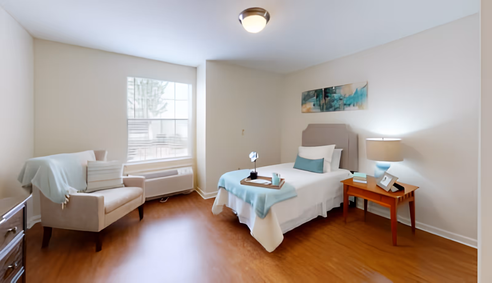 A bright and clean senior living bedroom with a single bed covered in white and light blue bedding. A wooden nightstand with a lamp, books, and a photo frame is beside the bed. There is a beige armchair with a light blue throw blanket and pillows near a large window with white blinds. The room has light-colored walls and wooden flooring, with a piece of abstract artwork hanging above the bed.