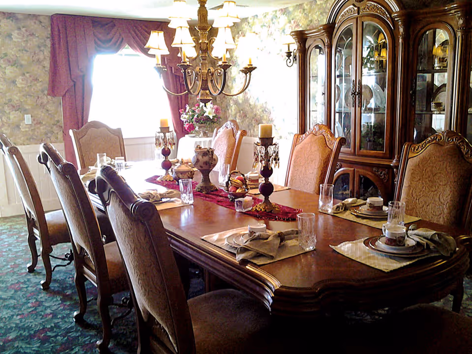 A formal dining room with a long wooden table set for six people. The table has placemats, napkins, glassware, and china cups. There are ornate chairs around the table, a chandelier hanging above, floral wallpaper, a window with burgundy curtains, and a large wooden china cabinet displaying dishes.