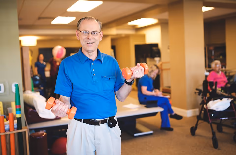 An elderly man wearing glasses and a blue shirt is smiling and holding orange dumbbells in both hands in a well-lit room. In the background, other seniors and a caregiver are visible, with one person sitting on a bench and another using a walker. The setting appears to be a rehabilitation or exercise area within a senior living facility.